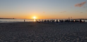 Una playa con filas de sillas y sombrillas, una persona caminando, y una puesta de sol al fondo.