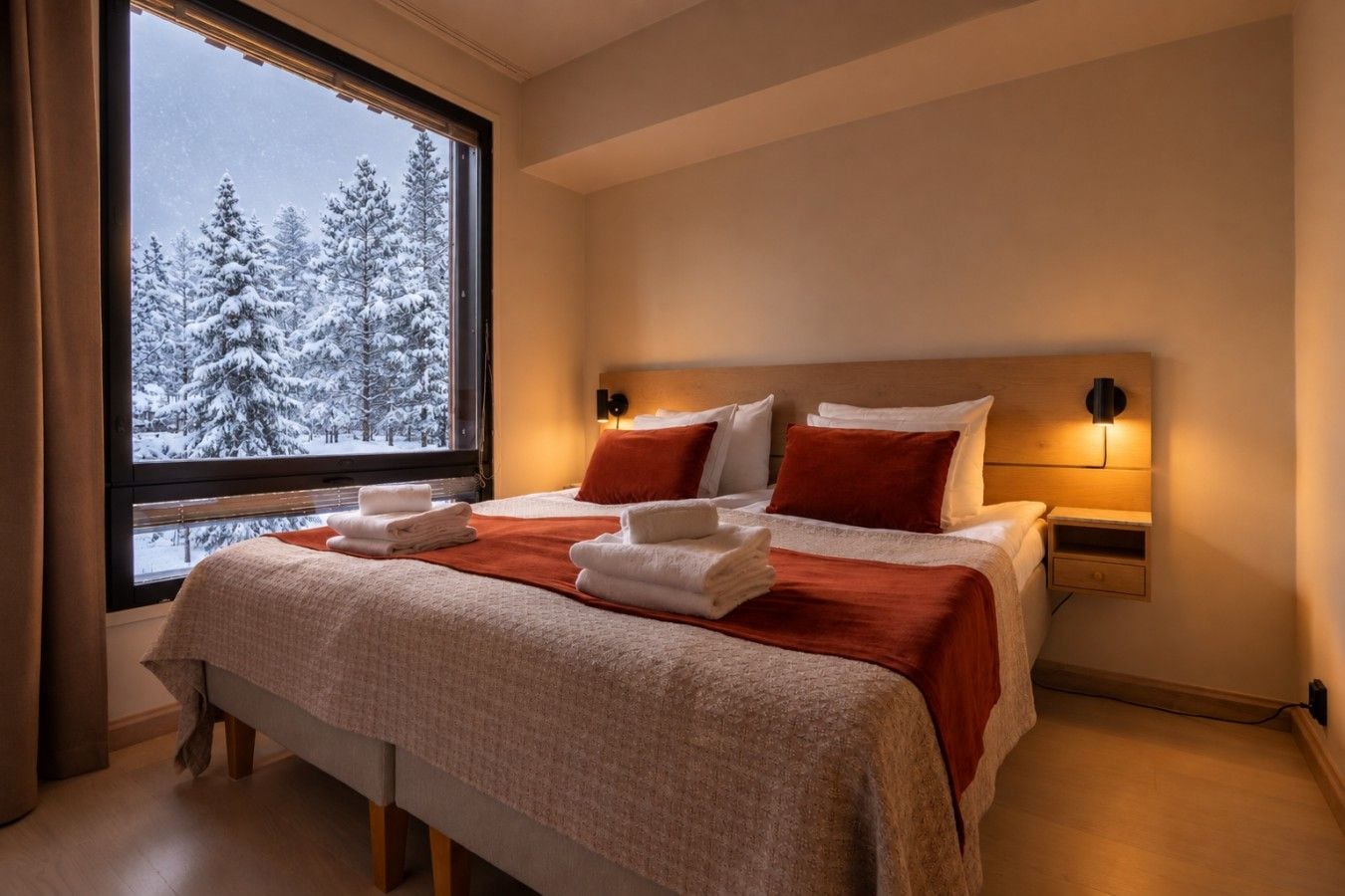 Une chambre d’hôtel avec un lit, deux tables de chevet, une fenêtre donnant sur des arbres couverts de neige et une petite table de chevet avec une lampe.