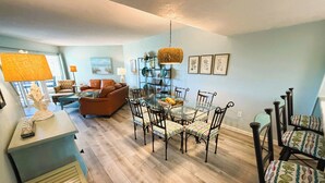 A living room with a brown sofa, a glass dining table with chairs, and a bookshelf. - Dove's Island Nest - Cozy Condo, Pool, Near Beach, Tennis Courts, Ocean Views (St. Augustine)