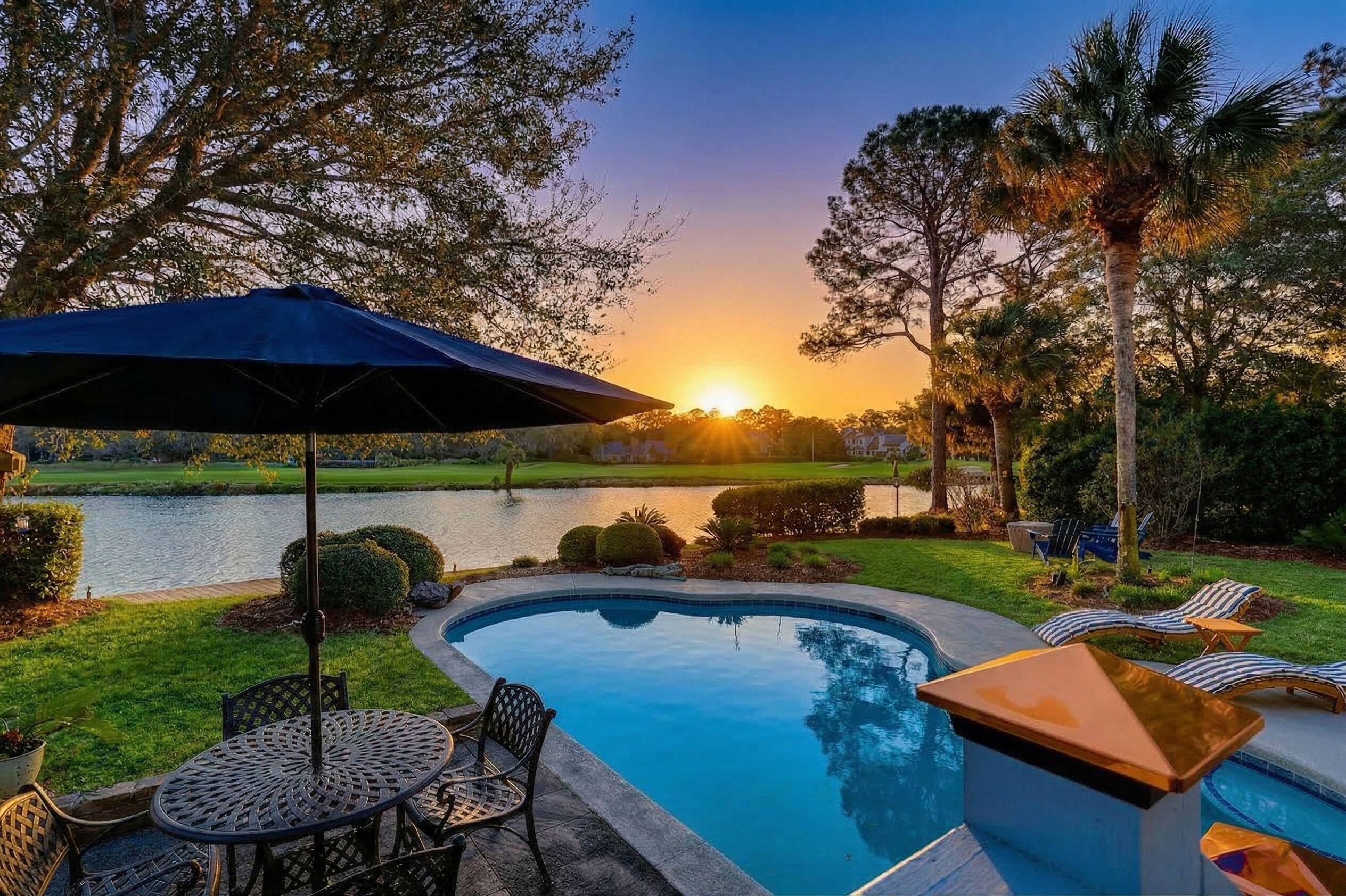 A swimming pool with lounge chairs and a table, surrounded by trees and a view of a lake at sunset.