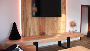 A modern living room with a wooden wall, a flat-screen TV mounted above a wooden bench, and a small table with decorative items. - Vacation in a half-timbered house, experience a real piece of living history. (Sehmatal-Neudorf)