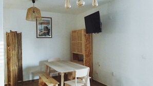 Ein Zimmer mit Holztisch, weißen Stühlen, einer Pendelleuchte, einem an der Wand befestigten Fernseher und einem Holzschrank.