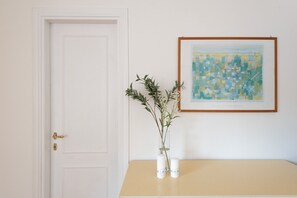 Une porte blanche avec une poignée dorée, un tableau abstrait encadré et un vase avec une plante sur une table de couleur beige.