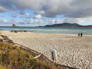A sandy beach with a castle-like structure in the distance, a clear sky, and a few people walking. - Charming Sea Villas-villa Sleeps 6IN Shared Pool (Stintino)