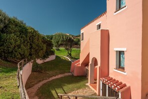 A building with a pink facade, a stone pathway, and a view of the surrounding greenery and hills. - Charming Sea Villas-villa Sleeps 6IN Shared Pool (Stintino)