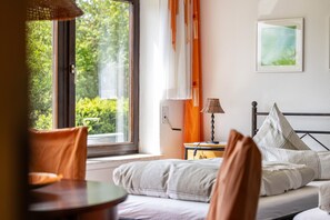 Ein Schlafzimmer mit Bett, Nachttisch, Lampe, einem Fenster mit Vorhängen und Blick ins Grüne.