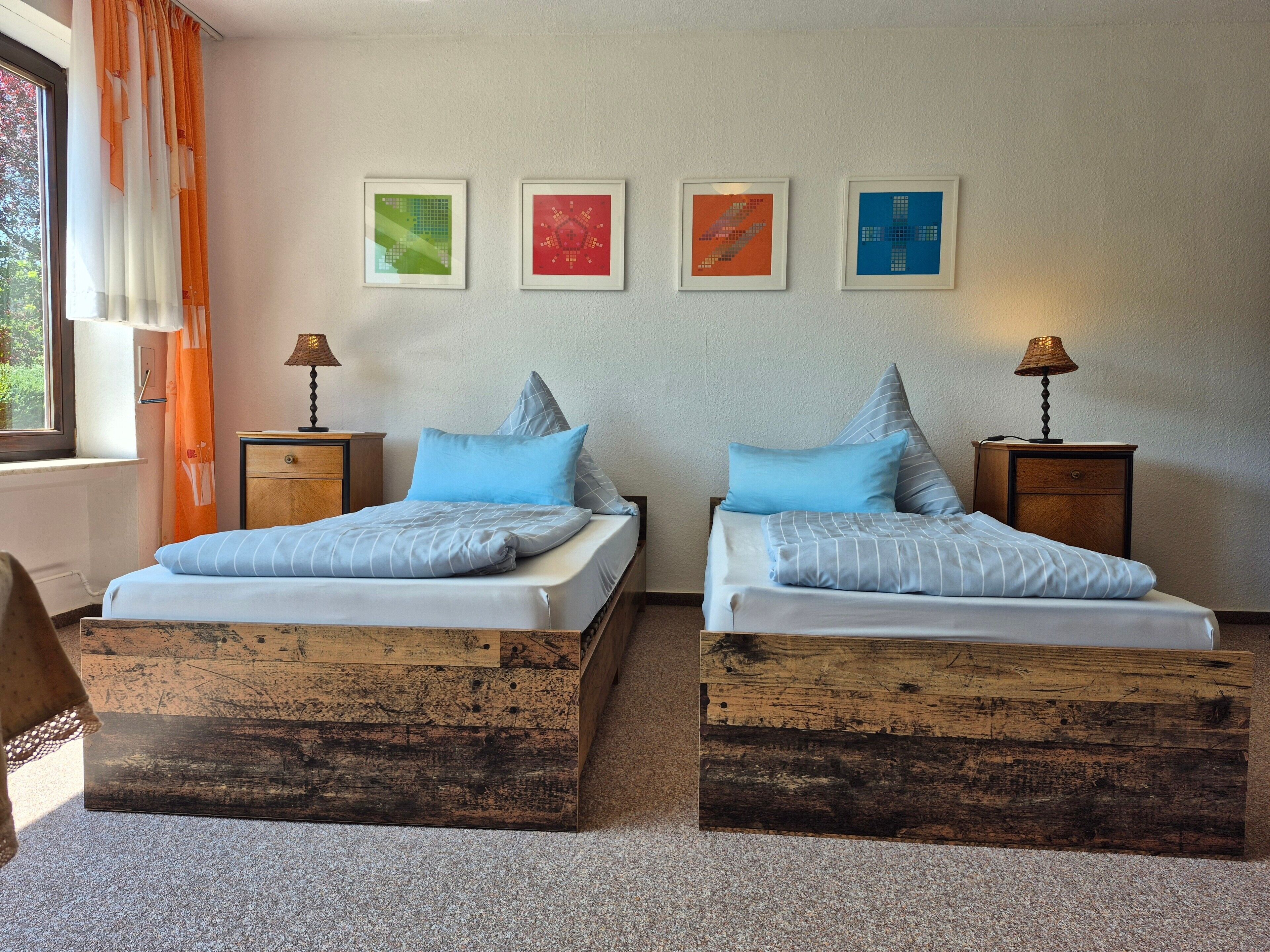 Une chambre avec deux lits, chacun doté d’une tête de lit en bois, deux tables de chevet avec des lampes, et trois œuvres d’art encadrées accrochées au mur.