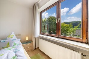 Ein Schlafzimmer mit großem Fenster, einem Bett mit weißer und grüner Decke, einem Nachttisch mit Lampe und einem Heizkörper.