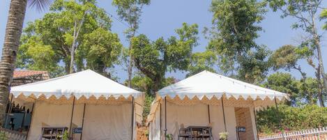 Two white tents with beige trim, each with a table and chairs, surrounded by greenery and trees.