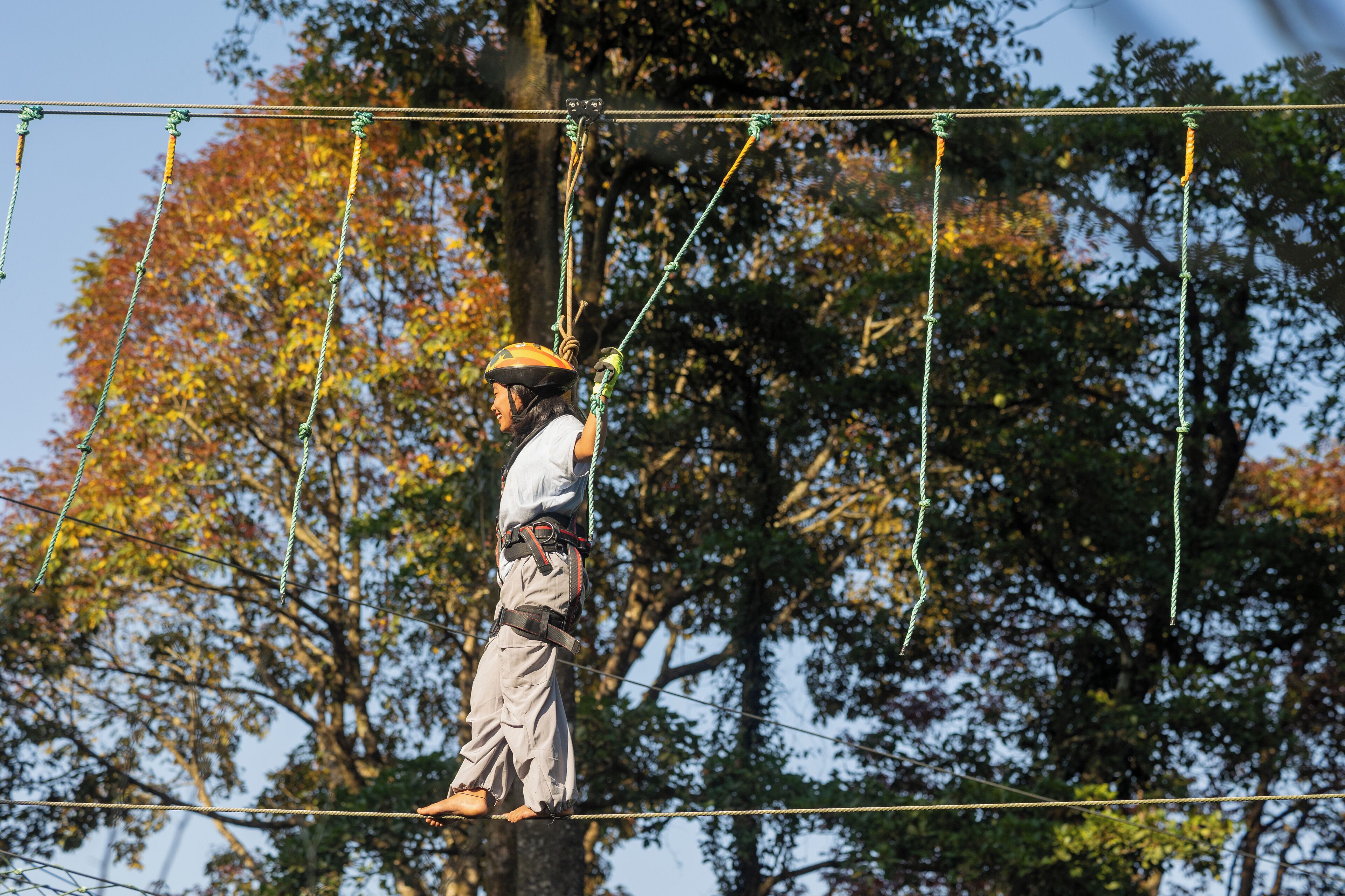 A person on a rope course with safety harnesses and helmets, surrounded by trees.