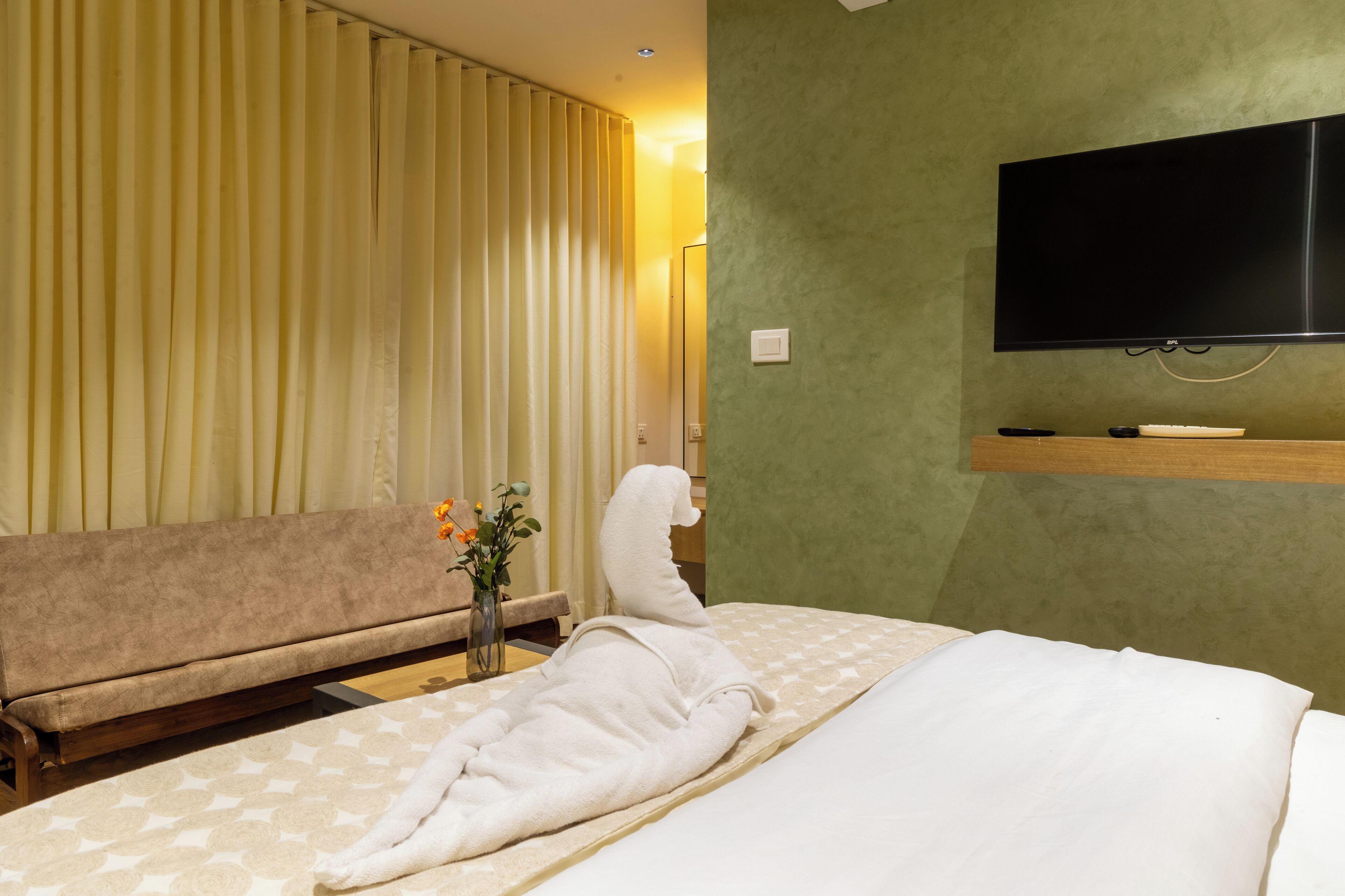 A hotel room with a bed, a sofa, a flat-screen TV, and a vase with flowers.