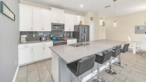 A modern kitchen with white cabinets, a large island with bar stools, a microwave, refrigerator, and a coffee maker. - Beautiful Paradise Palms Retreat 4 Bedroom Townhouse (Kissimmee)