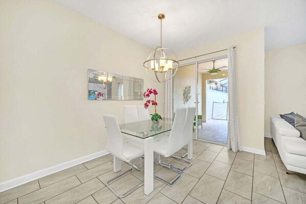 A dining area with a glass table and white chairs, a chandelier, and a large mirror on the wall. - Beautiful Paradise Palms Retreat 4 Bedroom Townhouse (Kissimmee)