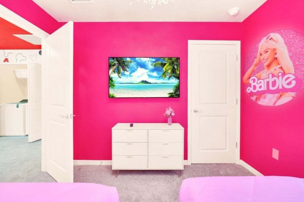 A room with a pink wall, a white dresser, a TV mounted on the wall, and a Barbie poster.