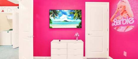 A room with a pink wall, a white dresser, a TV mounted on the wall, and a Barbie poster.