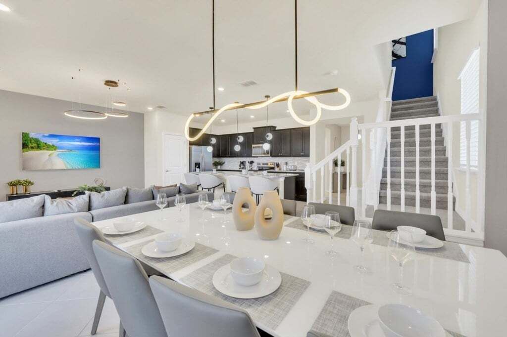 Ein moderner Essbereich mit einem weißen Tisch, gedeckt für sechs Personen, umgeben von grauen Sofas. Im Hintergrund ist eine Küche mit dunklen Schränken und einer weißen Arbeitsplatte erkennbar.
