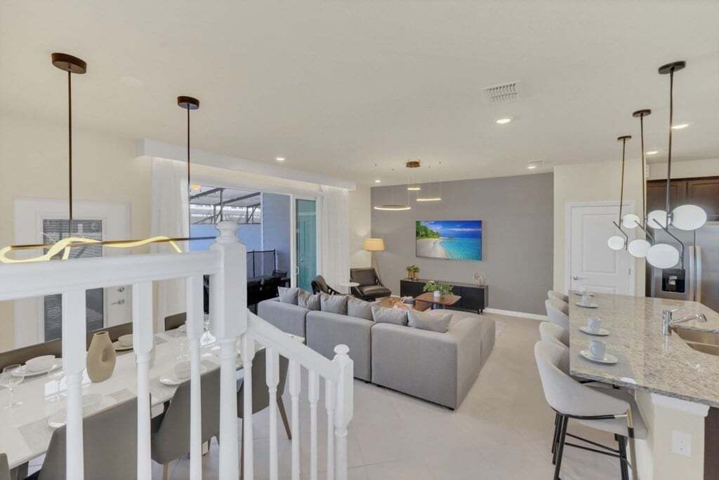 A modern living room with a grey sofa, a dining area with a white table, and a kitchen with a granite countertop.