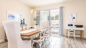 Un coin repas avec une table en bois, des chaises blanches et un lustre. Une porte vitrée donne sur l’extérieur.