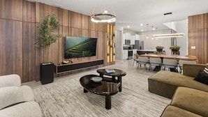 Ein modernes Wohnzimmer mit einem großen Flachbildfernseher, Wandverkleidung aus Holz, einem Essbereich mit Holztisch und -stühlen sowie einer Küche im Hintergrund.