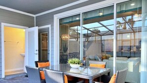 Une terrasse avec une table en verre et des chaises en osier, une plante en pot, et une vue sur l’intérieur à travers de grandes portes vitrées.