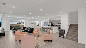 A spacious living area with a black sectional sofa, two pink armchairs, and a coffee table. A kitchen with a bar and a staircase is visible in the background. - Everyone Can Come So Much Space Pool Home! 15 Bedroom Villa (Kissimmee)