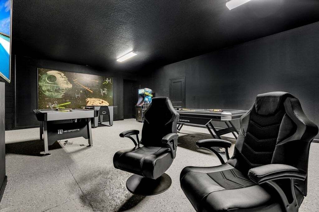 Sala de jogos com duas cadeiras gamer pretas, mesa de bilhar e uma tela grande exibindo um jogo.