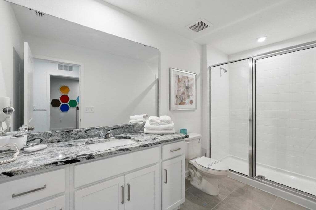 A modern bathroom with a large mirror, white cabinets, a marble countertop, a shower with a glass door, and a toilet.