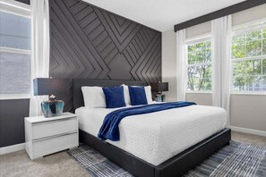 Un dormitorio moderno con una cama grande, cabecero oscuro, mesitas de noche blancas y vistas a zonas verdes a través de amplios ventanales.