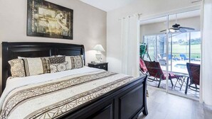 A bedroom with a large bed, a nightstand, a lamp, and a painting on the wall. There is a sliding glass door leading to an outdoor area with a pool and chairs. - 4 Bed Pool Home, Pool Table & More 4 Bedroom Villa (Davenport)