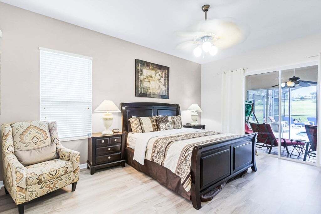 A bedroom with a bed, nightstands, a chair, a ceiling fan, and a view of the outdoors.