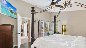 Una camera da letto con un grande letto, una ventola a soffitto, un lampadario, un quadro raffigurante una spiaggia e vista su un patio con posti a sedere all'aperto.