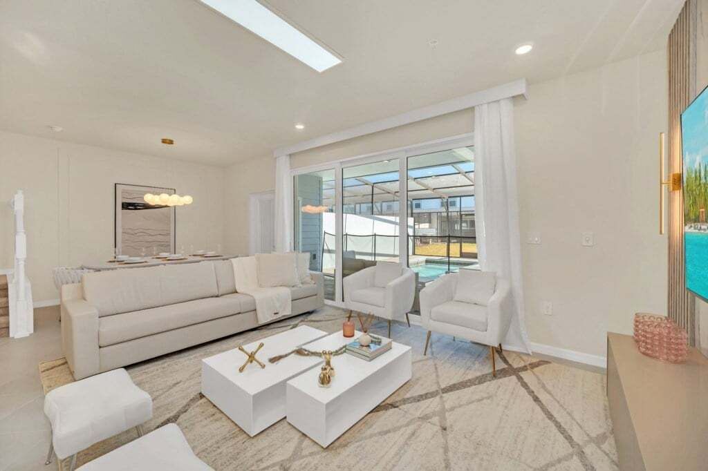 Un salon moderne avec un grand canapé, des fauteuils blancs et une table basse. Une grande fenêtre offre une vue sur une piscine et un balcon.