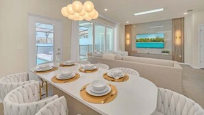 A modern dining area with a white table set for six, white upholstered chairs, and a large window offering a view of the outdoors. - Luxury Home With Splash Pool 5 Bed Townhome! 5 Bedroom Townhouse (Clermont)