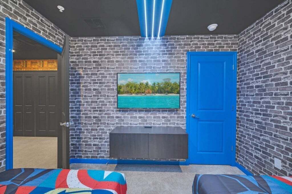 A room with a brick wall, a blue door, a television mounted on the wall, and a bench.