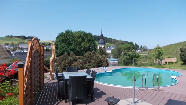 A pool area with a wooden deck, outdoor furniture, and a view of a hill with a church.