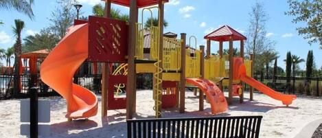A playground with slides and climbing structures, surrounded by sand and trees.