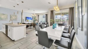 Una cucina moderna con isola centrale, un'area pranzo con un tavolo lungo e sedie, e un soggiorno con TV e posti a sedere.
