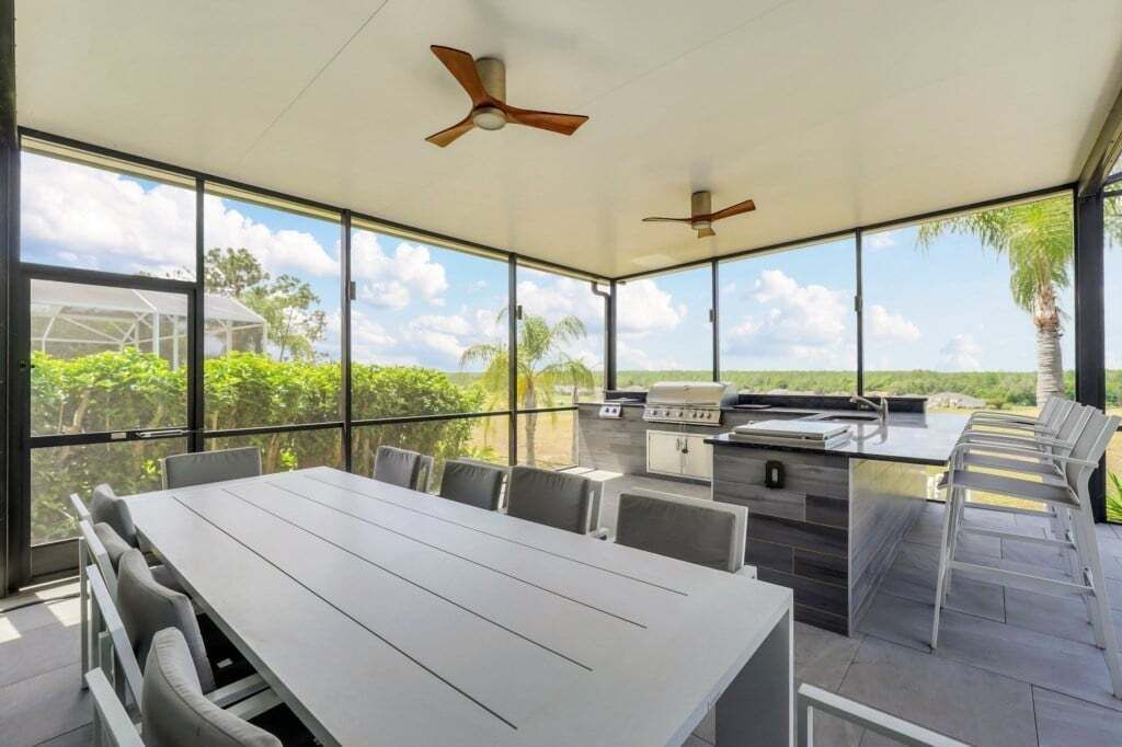 Un espace extérieur abrité, aménagé pour les repas, avec une table et des chaises, un barbecue et de grandes fenêtres offrant une vue sur l’extérieur.