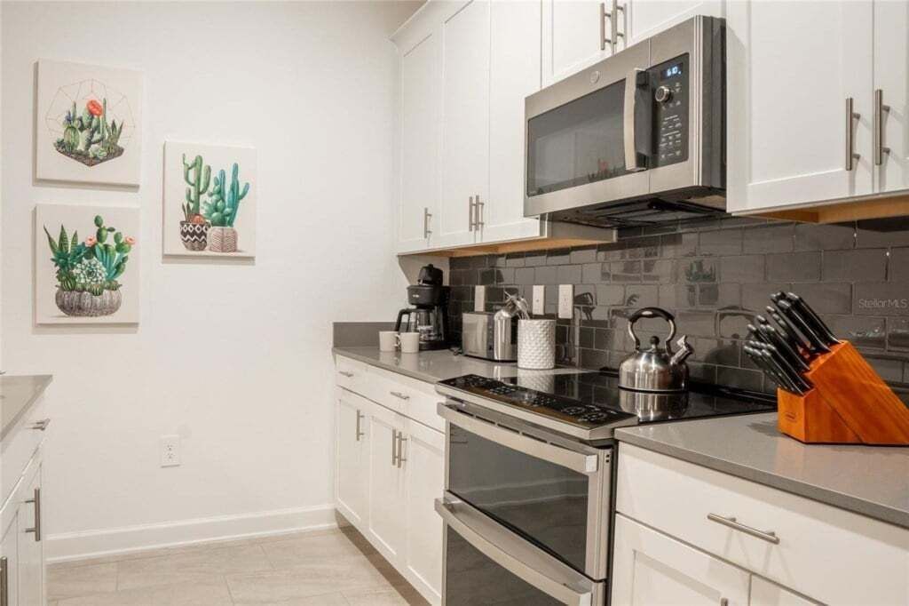 A modern kitchen with white cabinets, stainless steel appliances, and a backsplash. There are three framed botanical prints on the wall.