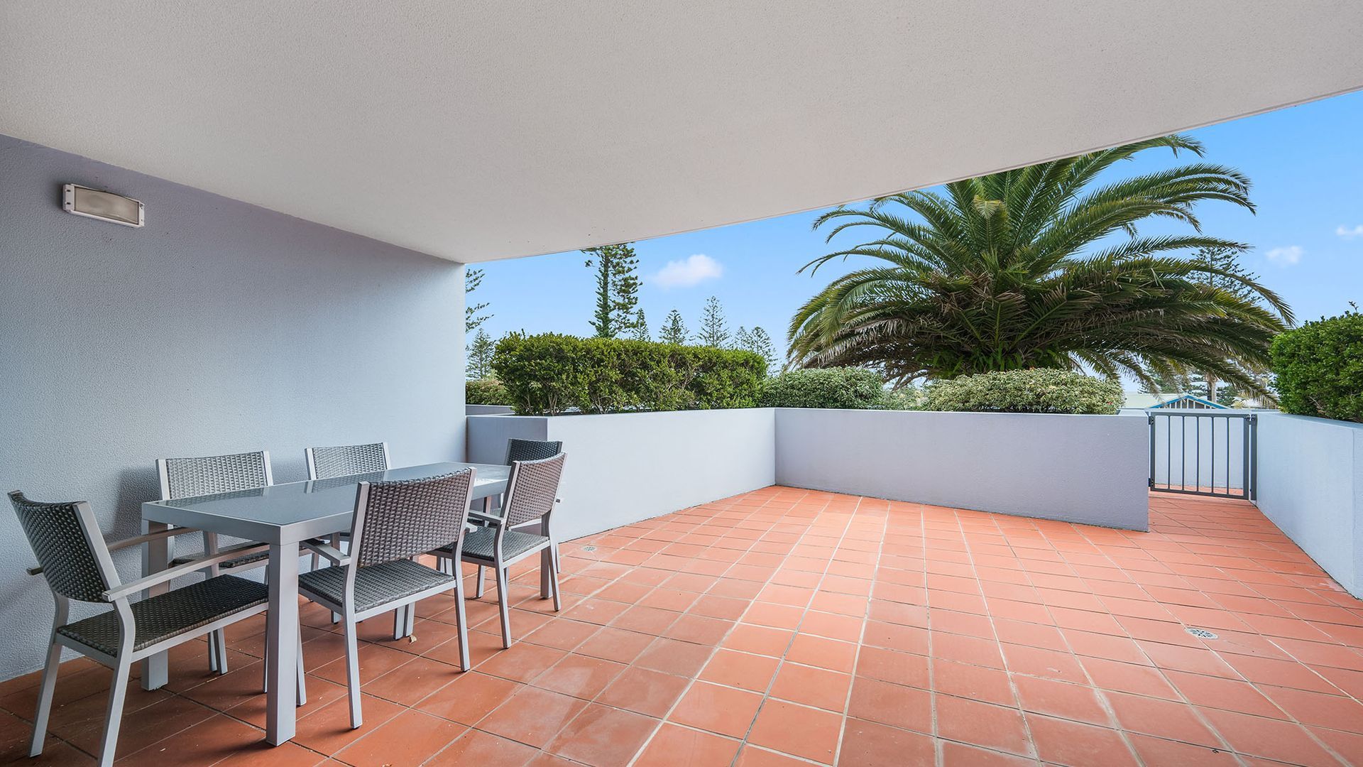 An outdoor patio with a table and chairs, surrounded by a low wall and a view of palm trees and greenery.