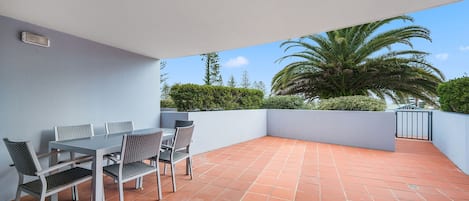 An outdoor patio with a table and chairs, surrounded by a low wall and a view of palm trees and greenery.