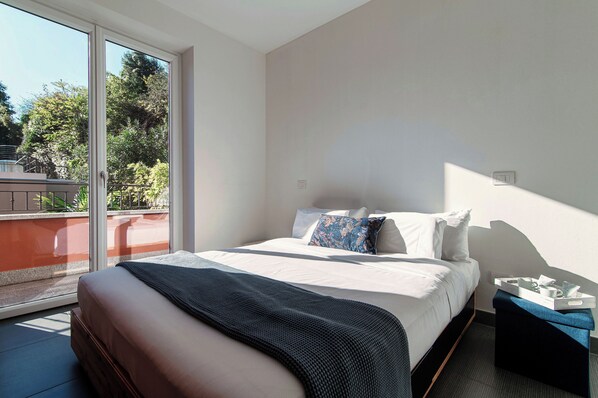 A bedroom with a bed, pillows, a bedside table, and a view of the outdoors through a sliding door. - Rosso Sunset (Colonno)