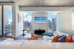 A modern living room with a white sofa, colorful cushions, a glass coffee table, and a painting of a beach. - Skyline Studio with Rooftop Pool & Parking (Miami)