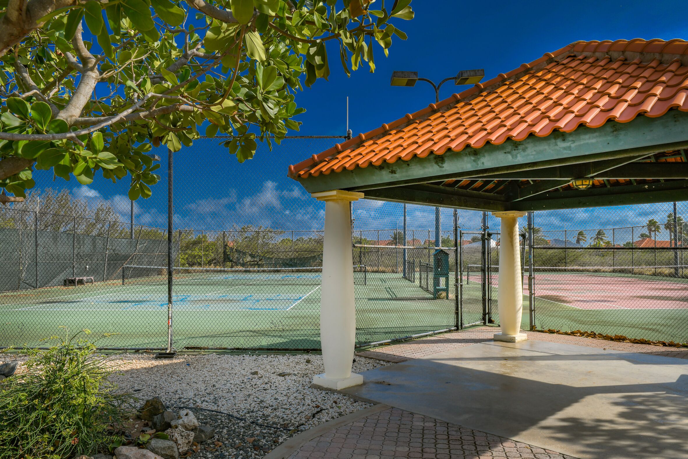 Un campo da tennis con un'area salotto coperta, circondato da una recinzione e alberi.