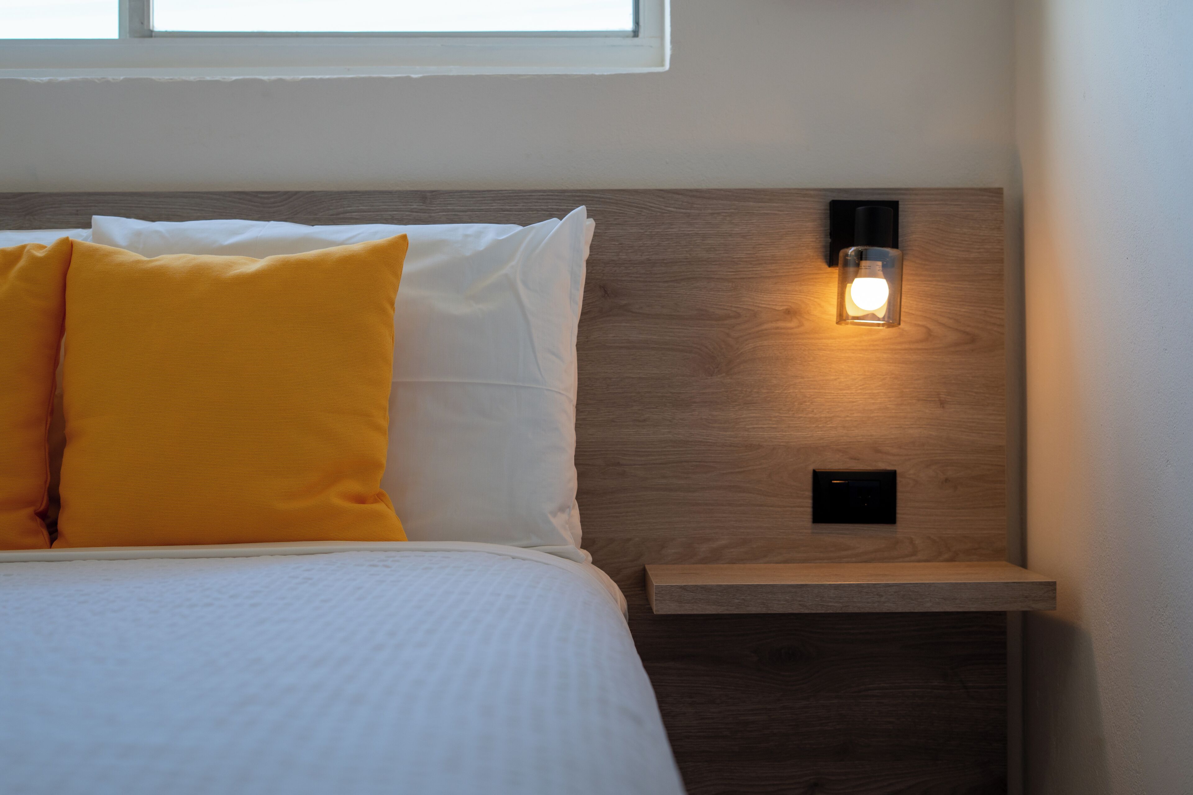 A neatly made bed with white and yellow pillows, a wooden headboard, and a bedside table with a wall-mounted light.