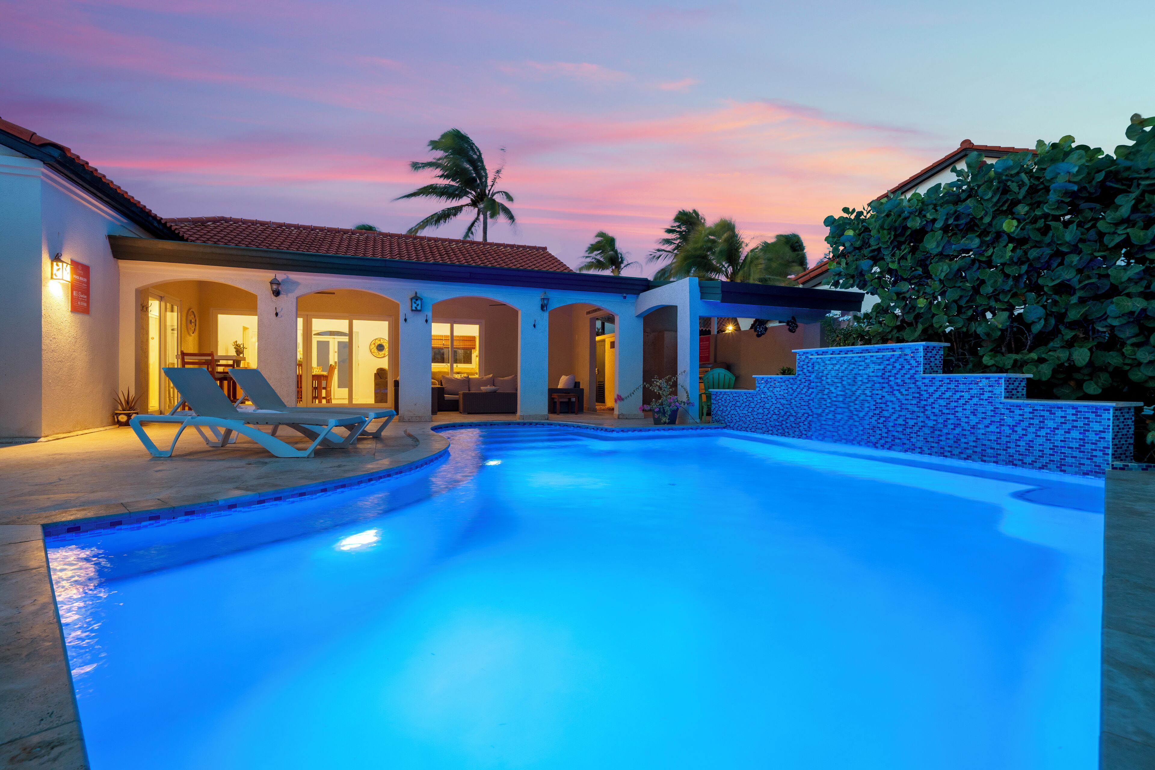 A swimming pool with lounge chairs, a covered patio, and a house with a tiled roof.
