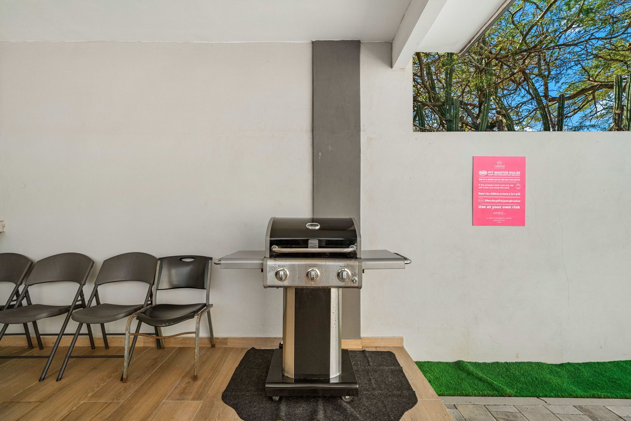 Uma área de churrasco moderna ao ar livre, com uma churrasqueira de aço inoxidável, três cadeiras dobráveis e uma placa de segurança vermelha na parede.