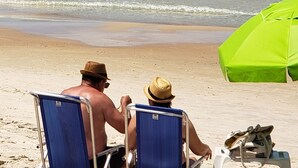 Two people sitting on beach chairs facing the ocean, with a green umbrella and a bag on the sand. - LM64 Lobster House in Bombinhas for 15 people Share Save (Bombinhas)