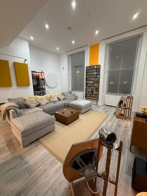 A modern living room with a sectional sofa, a coffee table, a bookshelf, and a large window with blinds. - Aberdeen Flat Union Street (Aberdeen)