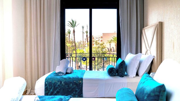 Ein Hotelzimmer mit großem Fenster, einem Bett mit blau-weißer Bettdecke und einem Balkon mit Blick auf Palmen und Gebäude.
